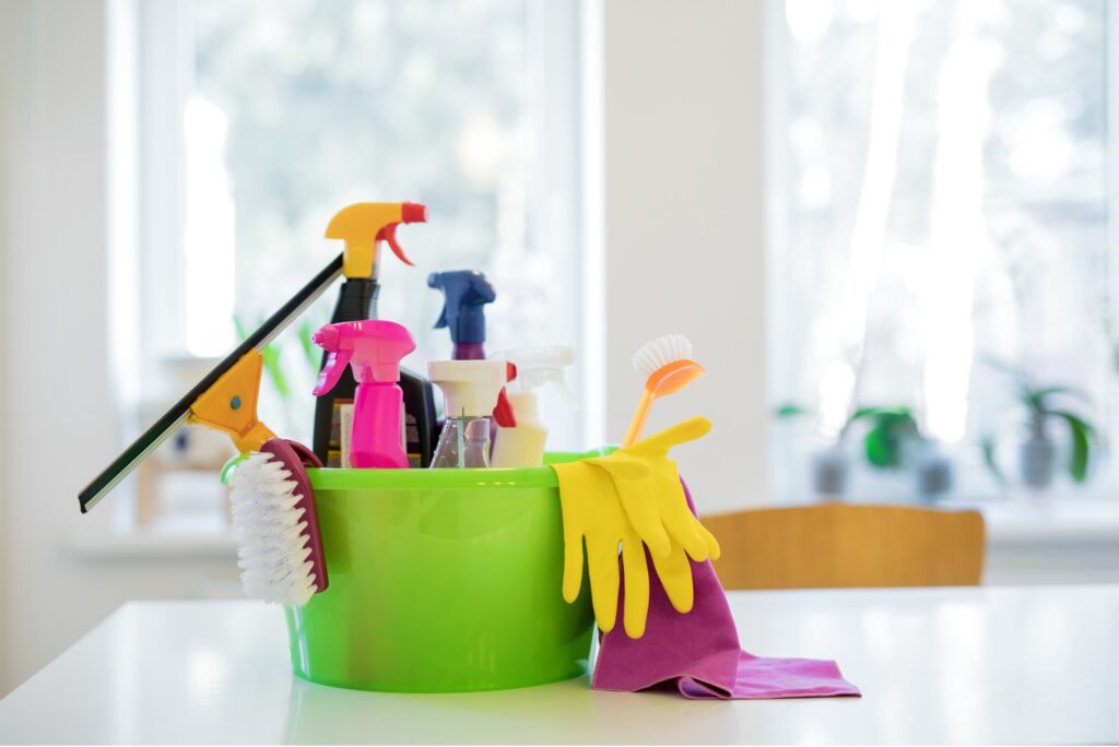 cleaning supply bucket with spray bottles, gloves, and brushes placed on a kitchen counter.